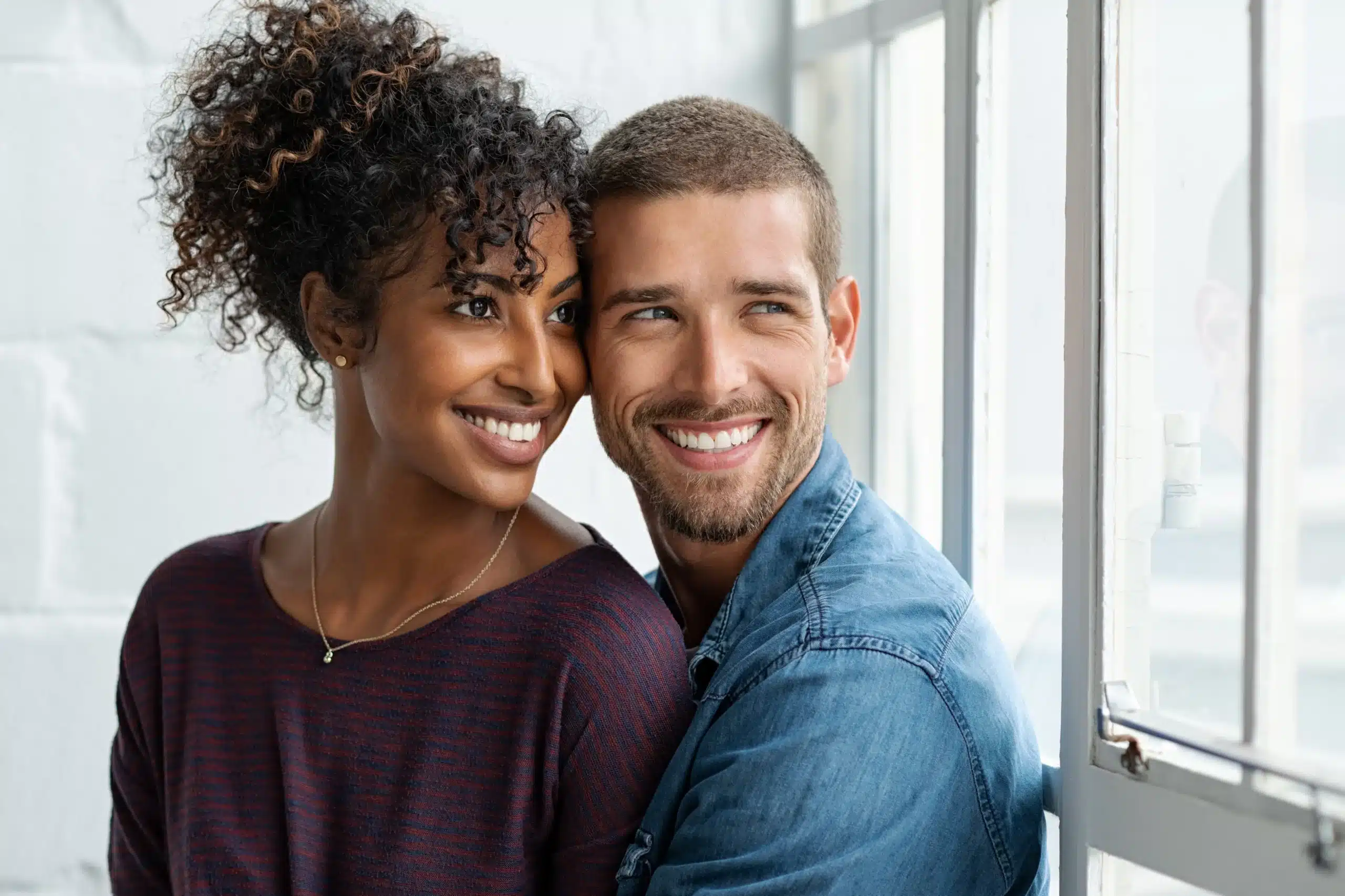 A beautiful, smiling couple embraces by a window, radiating happiness, a testament to Des Peres Dentistry.